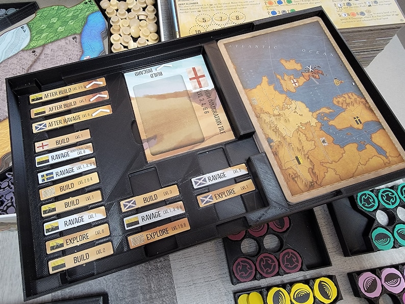 May include: A black board game tray with various coloured tokens and cards. The tray has slots for different game components, including a map, cards, and tokens. The tray is labelled with the words "Build", "Ravage", and "Explore".