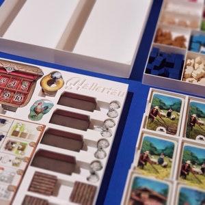 May include: A board game with various components, including cards, wooden blocks, and tokens, arranged in white trays. The game features a decorative chest with numbered slots and a rooster illustration. The word "Hellertau" is visible.