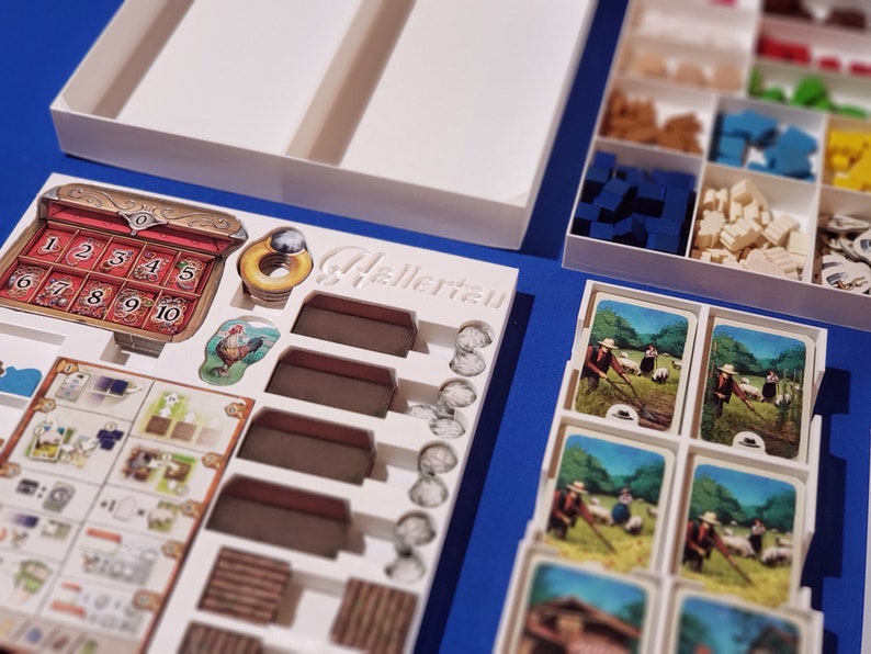 May include: A board game with various components, including cards, wooden blocks, and tokens, arranged in white trays. The game features a decorative chest with numbered slots and a rooster illustration. The word "Hellertau" is visible.