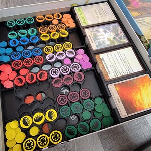 May include: A game organiser with compartments for storing game pieces and cards. The organiser has a black base with compartments for different coloured tokens, including yellow, red, green, blue, purple, and brown. The organiser also has compartments for cards, with the text "HEALTHY ISLAND" and "POWER BOOST FOR LIFE" visible.