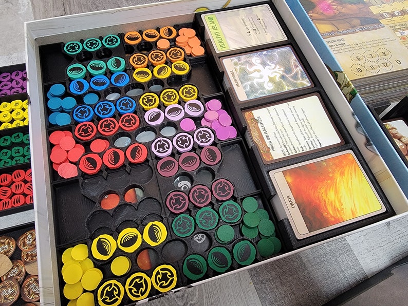 May include: A game organiser with compartments for storing game pieces and cards. The organiser has a black base with compartments for different coloured tokens, including yellow, red, green, blue, purple, and brown. The organiser also has compartments for cards, with the text "HEALTHY ISLAND" and "POWER BOOST FOR LIFE" visible.