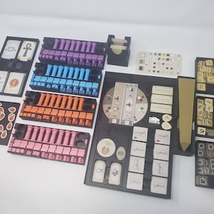 May include: A black organizer with compartments for storing game pieces and cards. The organizer includes compartments for different colored pieces, cards, and a wooden sword. The organizer is designed for the board game "The 7th Continent".