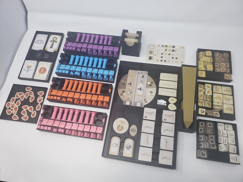 May include: A black organizer with compartments for storing game pieces and cards. The organizer includes compartments for different colored pieces, cards, and a wooden sword. The organizer is designed for the board game "The 7th Continent".