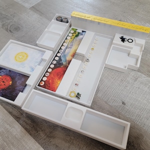 May include: White board game organizer with multiple compartments and trays. The organizer holds game pieces, cards with colorful artwork, and tokens. A yellow and gold decorative element is visible on the top.