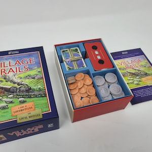 Puede incluir: Una caja azul con el texto "Village Rails" y una ilustración de un tren y ovejas. La caja contiene un juego con una bandeja roja y azul, una baraja de cartas y un conjunto de fichas.