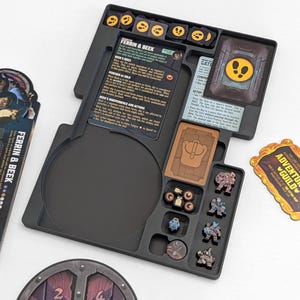 Może przedstawiać: Czarny organizer do gier planszowych z przegr&oacute;dkami na elementy gry, karty i kości. Organizer zawiera karty z tekstem, miniaturowe figurki i żetony. Tekst na kartach brzmi "FERRIN & BEEK" i "ADVENTURER'S GUILD".