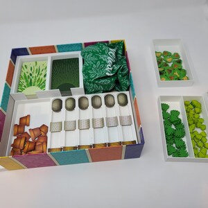 Puede incluir: Una caja blanca con una tapa a rayas de colores, que contiene varias piezas de juego. La caja tiene compartimentos para cartas, fichas y una bolsa de tela. Tambi&eacute;n hay cajas blancas separadas con fichas verdes y marrones.