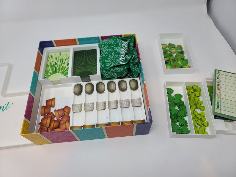 Puede incluir: Una caja blanca con una tapa a rayas de colores, que contiene varias piezas de juego. La caja tiene compartimentos para cartas, fichas y una bolsa de tela. Tambi&eacute;n hay cajas blancas separadas con fichas verdes y marrones.