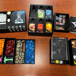 May include: A black organizer with multiple compartments for storing Catan: Starfarers game pieces. The organizer includes compartments for cards, dice, and small game pieces. The organizer is made of sturdy plastic and has a sleek, modern design.