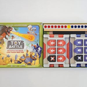 Puede incluir: Caja de juego de mesa abierta para "Toy Battle" con una ilustración vibrante de un dinosaurio y caballeros. La caja contiene piezas de juego rojas, azules y negras, junto con una pieza de madera con puntos rojos, amarillos y azules. Para 2+ jugadores.