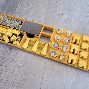 May include: A yellow board game organizer with various game pieces. The organizer has compartments for yellow buildings, bottles, and other game components. There are also gray game pieces and circular tokens with illustrations.