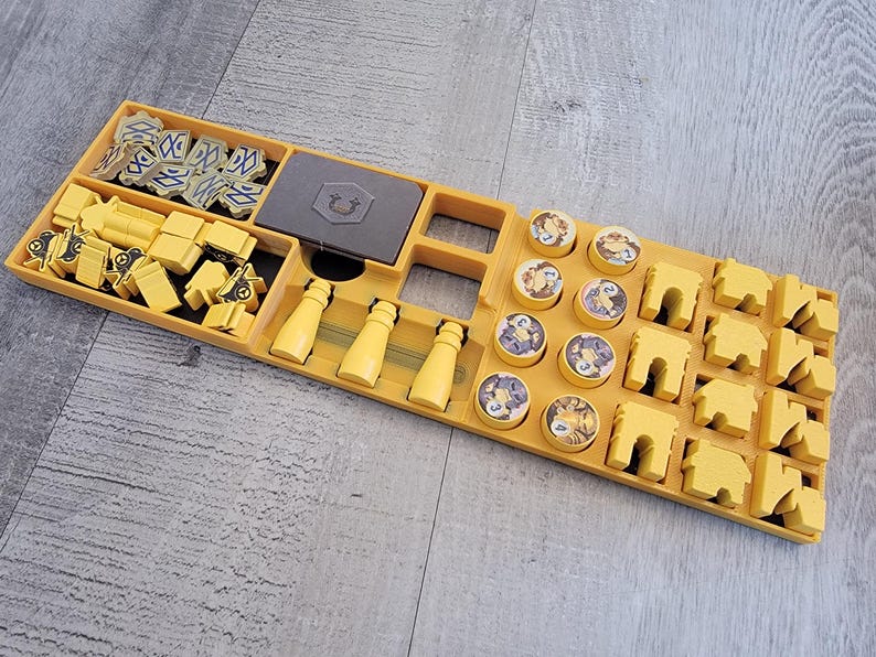May include: A yellow board game organizer with various game pieces. The organizer has compartments for yellow buildings, bottles, and other game components. There are also gray game pieces and circular tokens with illustrations.