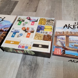 Könnte beinhalten: Ein Brettspiel-Set mit einer weißen Einlegeplatte und einer blau-weißen Schachtel mit dem Text "Iki Akebono Expansion Expansion". Das Spiel spielt in Japan und zeigt eine Brücke, Boote und Menschen in traditioneller Kleidung.