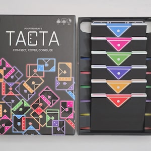 Puede incluir: Un juego de mesa titulado Tacta de Jason Tremblay, con una caja negra con ilustraciones de cartas geométricas de colores neón y el texto Connect, Cover, Conquer. El interior de la caja abierta contiene seis tarjeteros negros.