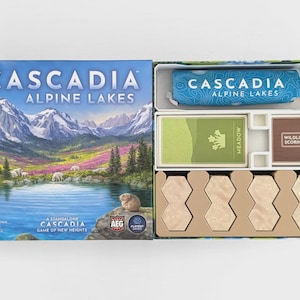 May include: The Cascadia Alpine Lakes board game box features a mountain lake landscape illustration. Inside, a blue fabric bag, a deck of green Meadow cards, a stack of brown Wildlife Scoring cards, and eight beige hexagonal tiles are organised.