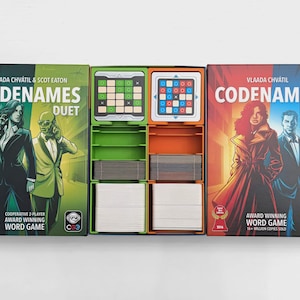 以下が含まれることがあります： Codenamesのボードゲームボックス2つ（Duetとオリジナル）が、開いたゲームオーガナイザーを挟んでいます。緑色のDuetボックスと赤色のオリジナルボックスには、2人の人物のイラストが描かれています。中央のトレイには、白とグレーのカードの束と2枚のグリッドタイルが収納されています。