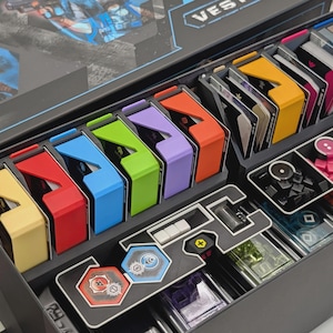 May include: A board game organizer box containing multiple colored plastic card holders in yellow, red, blue, green, purple, and orange. The box includes various compartments filled with decks of cards, small pink and brown cubes, and hexagonal tokens.