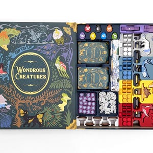 May include: Open board game box titled "Wondrous Creatures." The box is dark teal with gold accents and illustrations of fantastical creatures. Inside are game pieces in various colours, including cards, tokens, and player pieces.