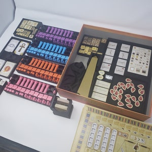 May include: A game organizer for the board game "The 7th Continent". The organizer includes multiple compartments for storing the game's components, including cards, tokens, and dice. The organizer is made of black plastic and has a brown cardboard base.