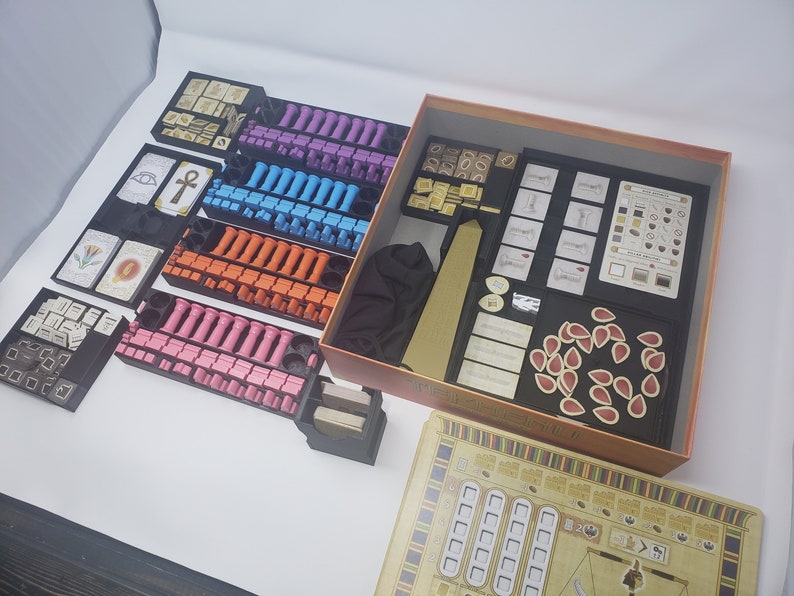 May include: A game organizer for the board game "The 7th Continent". The organizer includes multiple compartments for storing the game's components, including cards, tokens, and dice. The organizer is made of black plastic and has a brown cardboard base.