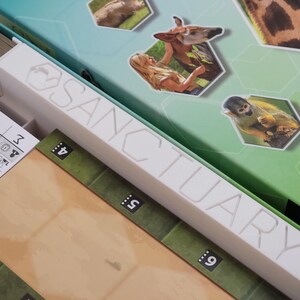 May include: A board game box with the word "SANCTUARY" in white lettering. The box is green and teal, with hexagonal images of animals. Inside are game components, including cards and a game board.