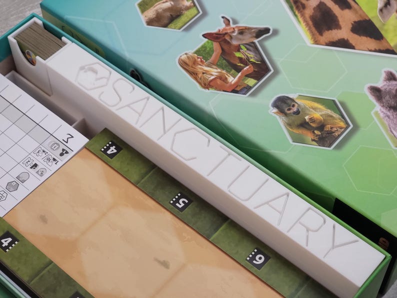May include: A board game box with the word "SANCTUARY" in white lettering. The box is green and teal, with hexagonal images of animals. Inside are game components, including cards and a game board.