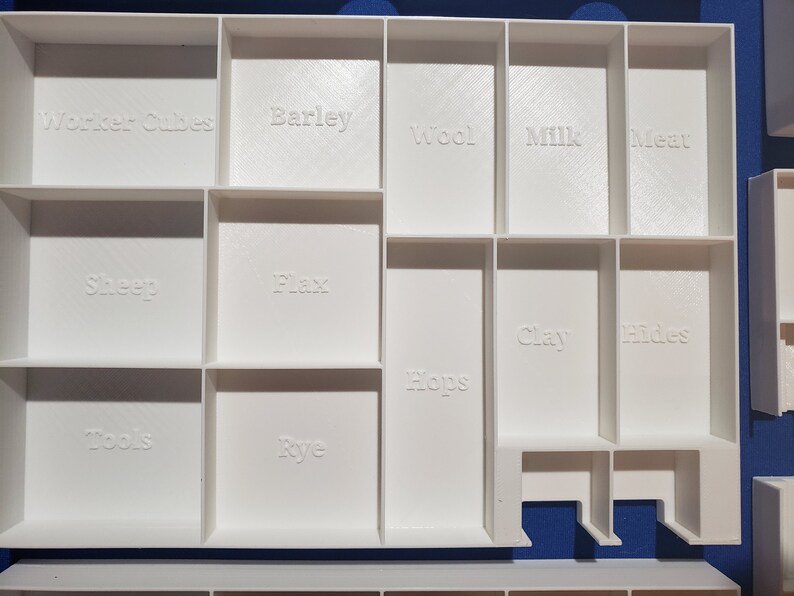 May include: A white, compartmentalized organizer with labeled sections for game components. The labels include "Worker Cubes," "Barley," "Wool," "Milk," "Meat," "Sheep," "Flax," "Hops," "Clay," "Hides," "Tools," and "Rye."