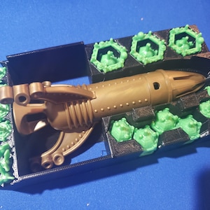 May include: A gold-colored plastic toy spaceship with a green plastic base. The spaceship has a pointed nose and a curved tail. The base has a series of hexagonal compartments.