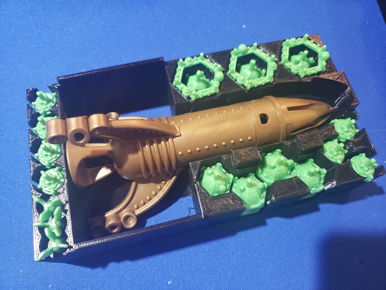 May include: A gold-colored plastic toy spaceship with a green plastic base. The spaceship has a pointed nose and a curved tail. The base has a series of hexagonal compartments.