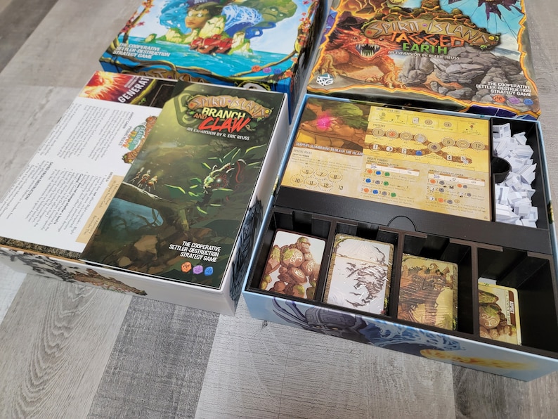 May include: A Spirit Island board game expansion called "Branch and Claw" with a green and brown box featuring a creature with a claw. The box is next to a Spirit Island game box with a blue and green box featuring a dragon. The game box is open and shows the game board and components.