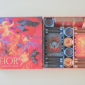 May include: Board game box titled "IGOR" with fantasy artwork on the lid. The box is open, revealing game components arranged in a pink tray. The components include cards, tokens, and other game pieces.