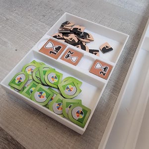 May include: A white, compartmentalized game organizer filled with game pieces. The pieces include green and multi-colored tokens, brown tiles with hourglass designs, and black and brown wooden pieces. The organizer is on a light gray surface.