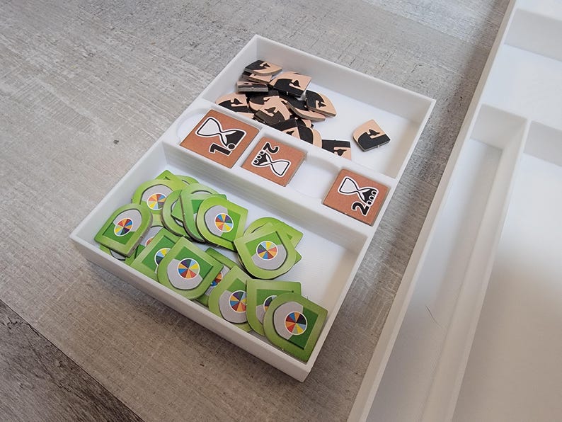 May include: A white, compartmentalized game organizer filled with game pieces. The pieces include green and multi-colored tokens, brown tiles with hourglass designs, and black and brown wooden pieces. The organizer is on a light gray surface.