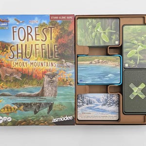 May include: The Forest Shuffle Smoky Mountains board game box features an illustration of an otter in a river with forest scenery. The open box contains a brown plastic insert holding stacks of game cards with nature-themed illustrations.