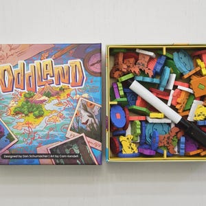 May include: The board game "Oddland" is displayed, with its box and contents visible. The box features a map design with the game's title in orange and blue. Inside, various colorful wooden game pieces and a white marker are neatly arranged.