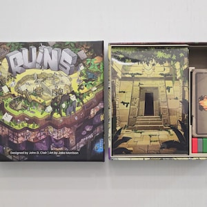 May include: Board game box set titled "RUINS" with colourful artwork depicting a jungle scene and ancient ruins. The open box reveals game components, including cards and coloured pieces. Designed by John D. Clair, art by Jake Morrison.