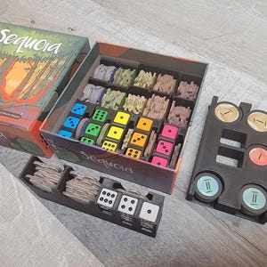 Puede incluir: Un inserto organizador de juego negro y gris para el juego de mesa "Sequoia". El inserto tiene compartimentos para guardar los componentes del juego, incluidos dados, fichas y cartas. El inserto está diseñado para caber dentro de la caja del juego.