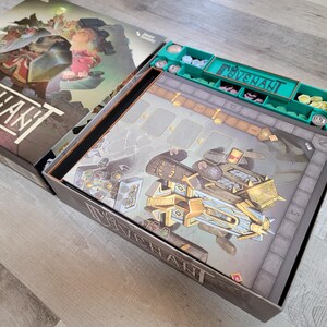 May include: Open board game box with the game title "Covenant" visible. The box contains a game board with a detailed illustration of a futuristic vehicle and a green organizer with game pieces. The box is on a light wood surface.