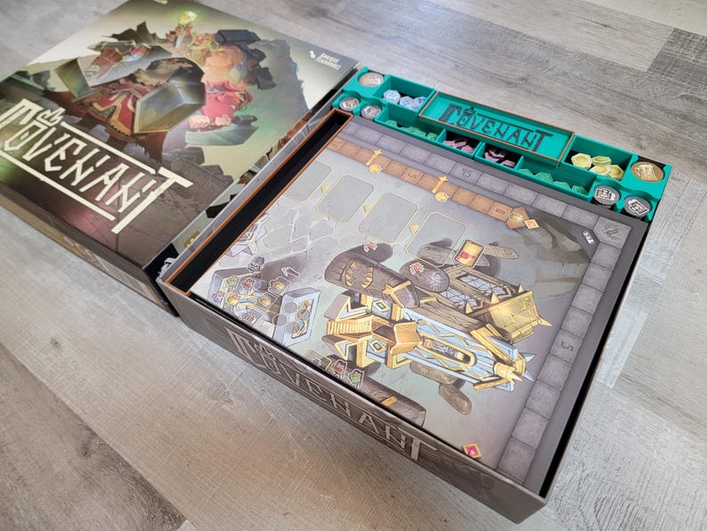 May include: Open board game box with the game title "Covenant" visible. The box contains a game board with a detailed illustration of a futuristic vehicle and a green organizer with game pieces. The box is on a light wood surface.