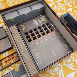 May include: Open board game box with compartments for cards, tokens, and player pieces. The game board features spaces for player tokens and game information. The box is brown with gray and gold accents, and the game is titled "Roll Player."