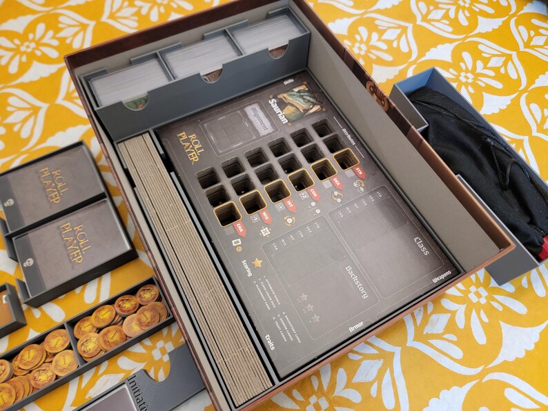 May include: Open board game box with compartments for cards, tokens, and player pieces. The game board features spaces for player tokens and game information. The box is brown with gray and gold accents, and the game is titled "Roll Player."