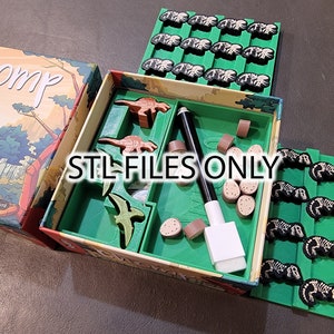 May include: A green game board with the word "Chomp" on the lid. The board has a green tray with dinosaur figurines, a white eraser, and a black marker. There are also several small brown pieces in the tray. The game board is open and shows the inside of the tray. There are also several green tiles with black dinosaur skeletons on them.