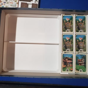 May include: A board game box with a white interior, divided into compartments. Several game cards with illustrations of houses and gates are visible. Other game pieces are in a separate compartment.