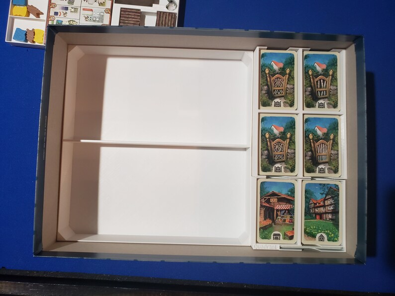 May include: A board game box with a white interior, divided into compartments. Several game cards with illustrations of houses and gates are visible. Other game pieces are in a separate compartment.