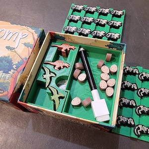 May include: A green box with the word "Chomp" on the lid. The box contains a dinosaur-themed board game with wooden pieces, a dry-erase marker, and an eraser. The game includes a green board with dinosaur figures and a separate tray with brown dinosaur bones.