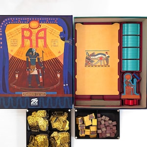 May include: Open board game box with the title "RA" in large letters. The box art features an Egyptian pharaoh and hieroglyphs. Inside are game pieces, including gold-colored tokens and teal colored game pieces.