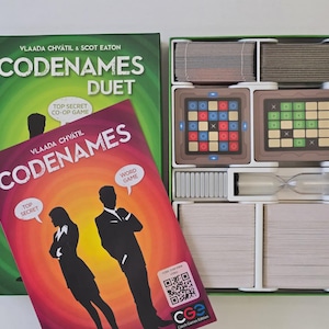 May include: A Codenames Duet board game set, featuring a green box and a red game box cover. The game includes cards, game boards, and an hourglass. The cover displays the game title and silhouettes of a man and woman.