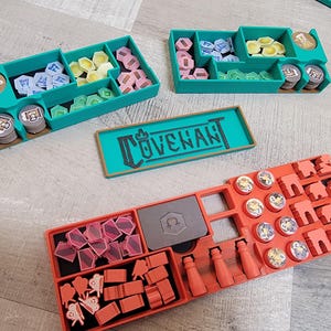 May include: A teal organizer with multiple compartments filled with game pieces in various colors, including pink, yellow, blue, and green. A rectangular teal tray with the word "COVENANT" is also present. An orange organizer is also visible.