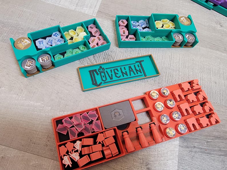 May include: A teal organizer with multiple compartments filled with game pieces in various colors, including pink, yellow, blue, and green. A rectangular teal tray with the word "COVENANT" is also present. An orange organizer is also visible.