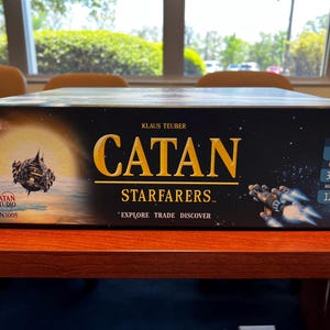 May include: A black box game with the title "CATAN STARFARERS" in gold lettering. The game is for 3-4 players, ages 14 and up, and takes 120 minutes to play.
